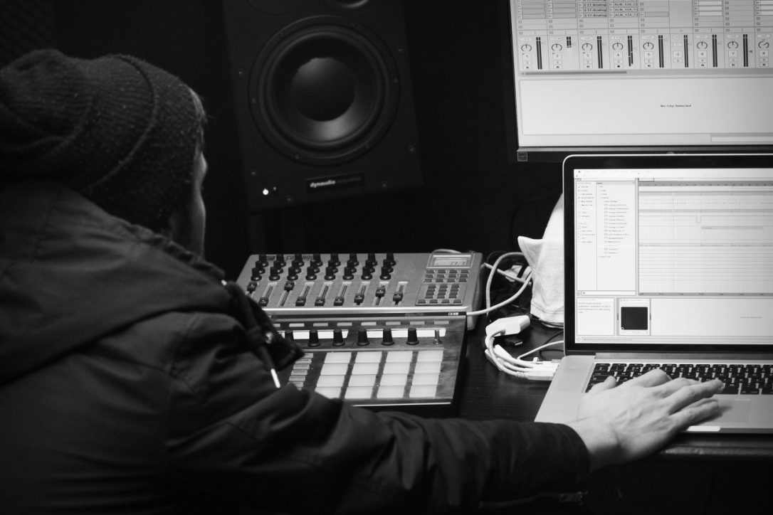 A producer at a studio desk with monitors, a laptop and a controller
